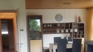 A spacious living area with a dining table and chairs, a kitchen area with cabinets and a clock, and a visible door leading to another room.