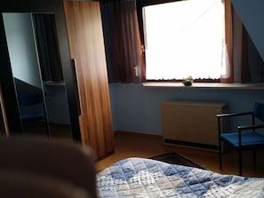 A bedroom with a bed, a desk, a chair, a radiator, and a window with curtains. - Aan de Frankenbeek (Sinzig)
