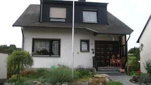 A two-story house with a dark roof, a front door with a small porch, and a garden with rocks and plants.
