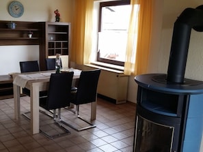 A dining area with a table, chairs, a clock, a window with curtains, and a wood stove. - Aan de Frankenbeek (Sinzig)