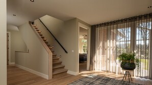 Un vaste espace de vie agrémenté d’un escalier en bois, d’une grande fenêtre avec des rideaux légers et d’une plante en pot posée sur un support.