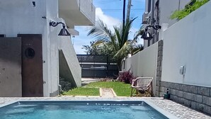 Una piscina con agua azul cristalina, rodeada por un edificio blanco con una verja negra, un pequeño jardín con un banco y una palmera.