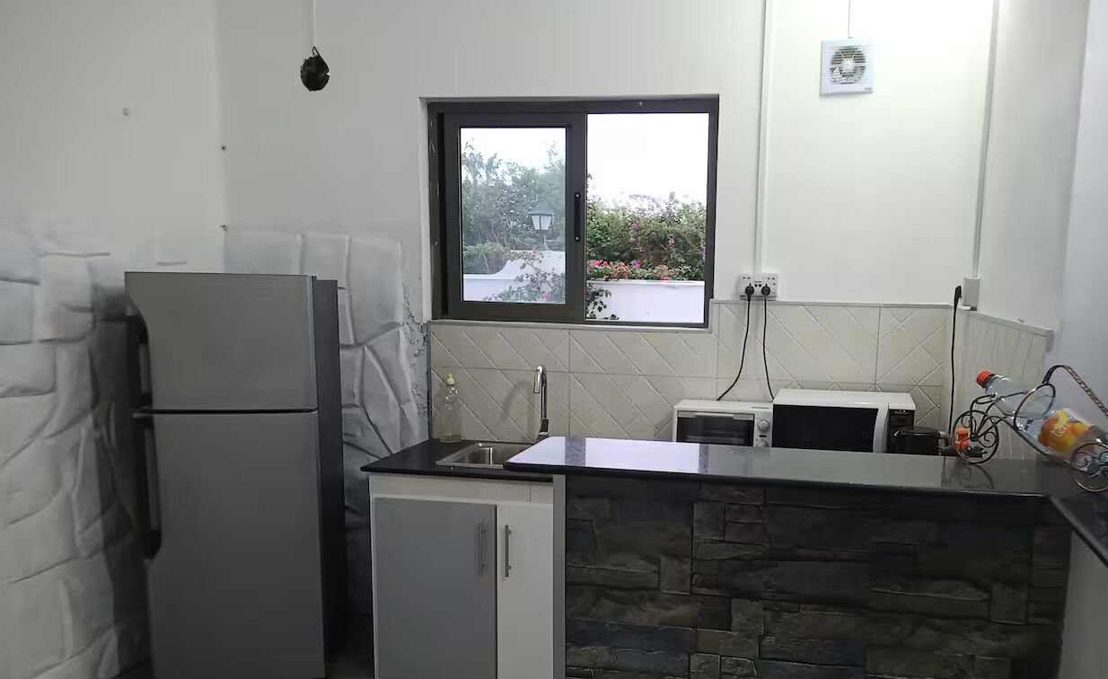 Cozinha compacta com geladeira, micro-ondas e pia. Há uma janela com vista para o verde lá fora.