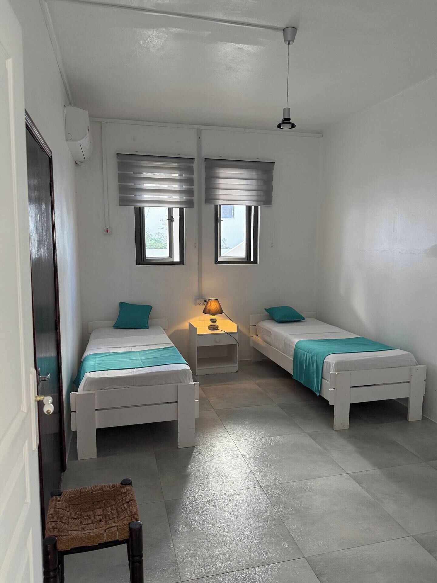 Une petite chambre propre, avec deux lits simples, une table de chevet avec une lampe et une fenêtre avec des stores.