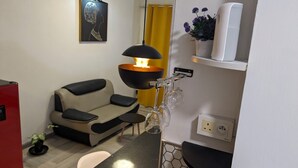 Un espacio habitable pequeño con sofá, lámpara colgante, mesita, planta en maceta y estante con un electrodoméstico blanco.