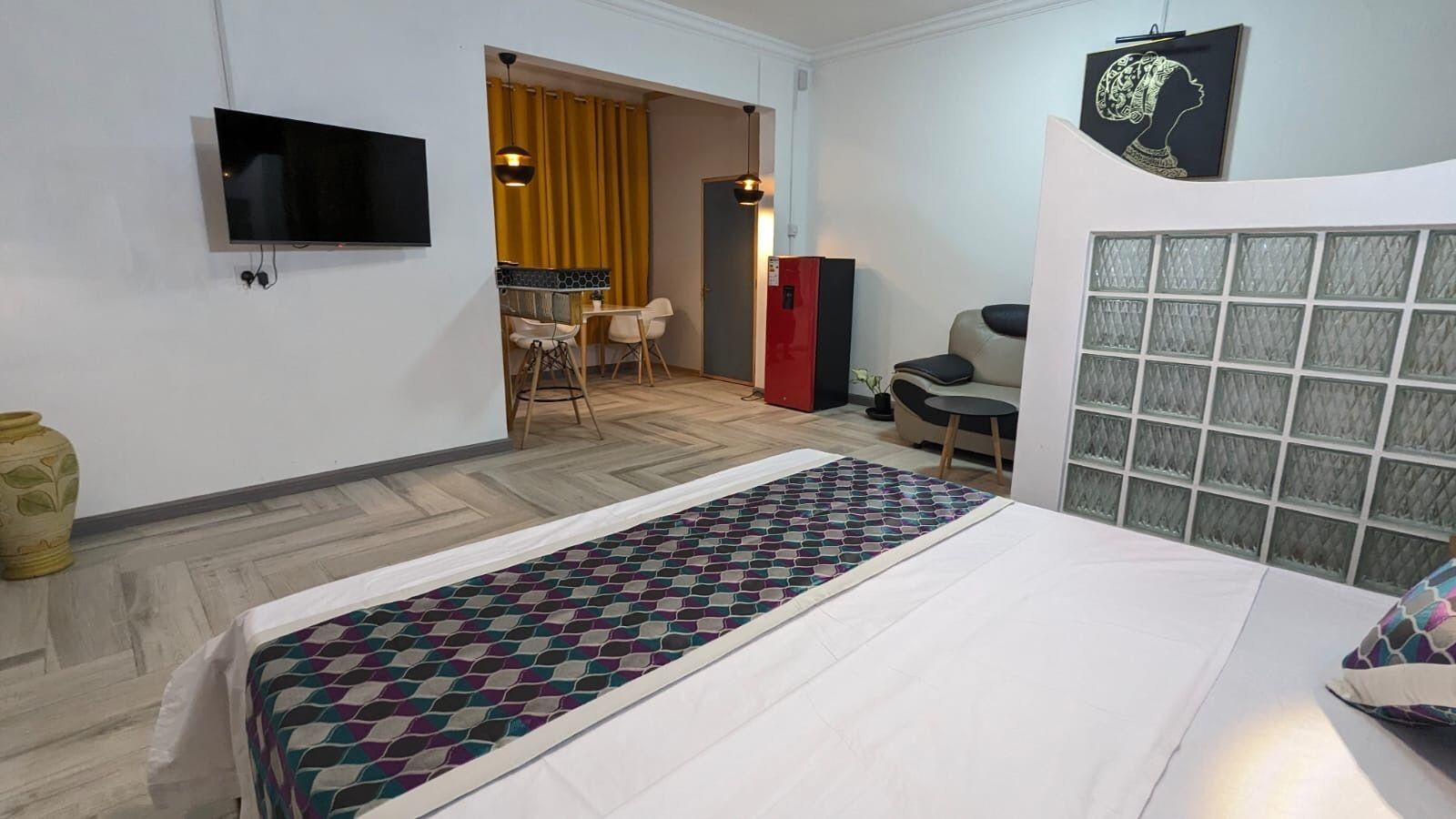 Une chambre d’hôtel avec un lit, un téléviseur fixé au mur, un petit coin repas avec des chaises, un réfrigérateur rouge et un panneau décoratif mural.