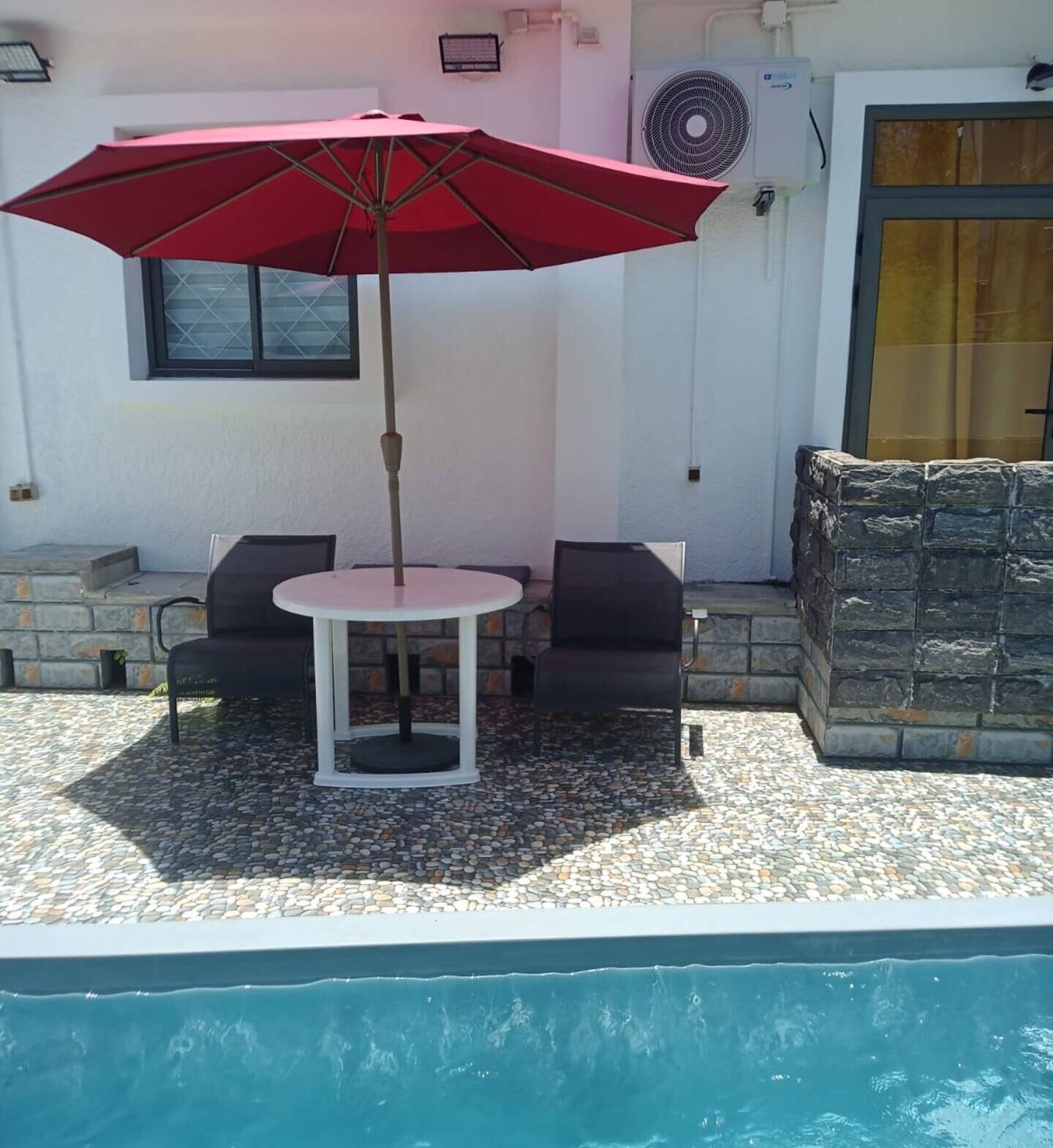 Een terras met een tafel, stoelen, een rode parasol en een zwembad.