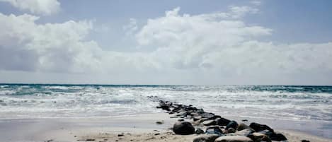 A beach with a row of rocks leading to the ocean, under a partly cloudy sky.
