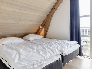 Ein Schlafzimmer mit schrägem Dach, zwei Betten mit weißer Bettwäsche, einem Holz-Kopfteil und Blick ins Freie durch ein Fenster.