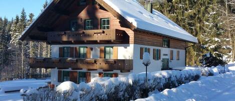 A two-story log cabin with a steeply pitched roof, surrounded by snow-covered trees and a clear sky.