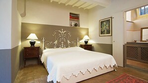 A bedroom with a large bed, two bedside tables with lamps, a wooden headboard, a red patterned rug, a small shelf with decorative items, and a door leading to another room. - Belvilla by OYO Villa Topazio REQ (Cortona)