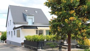 A two-story house with a grey tiled roof, a small balcony, and a fenced yard with trees and shrubs. - Holiday Apartment Festungsberg (Koblenz)