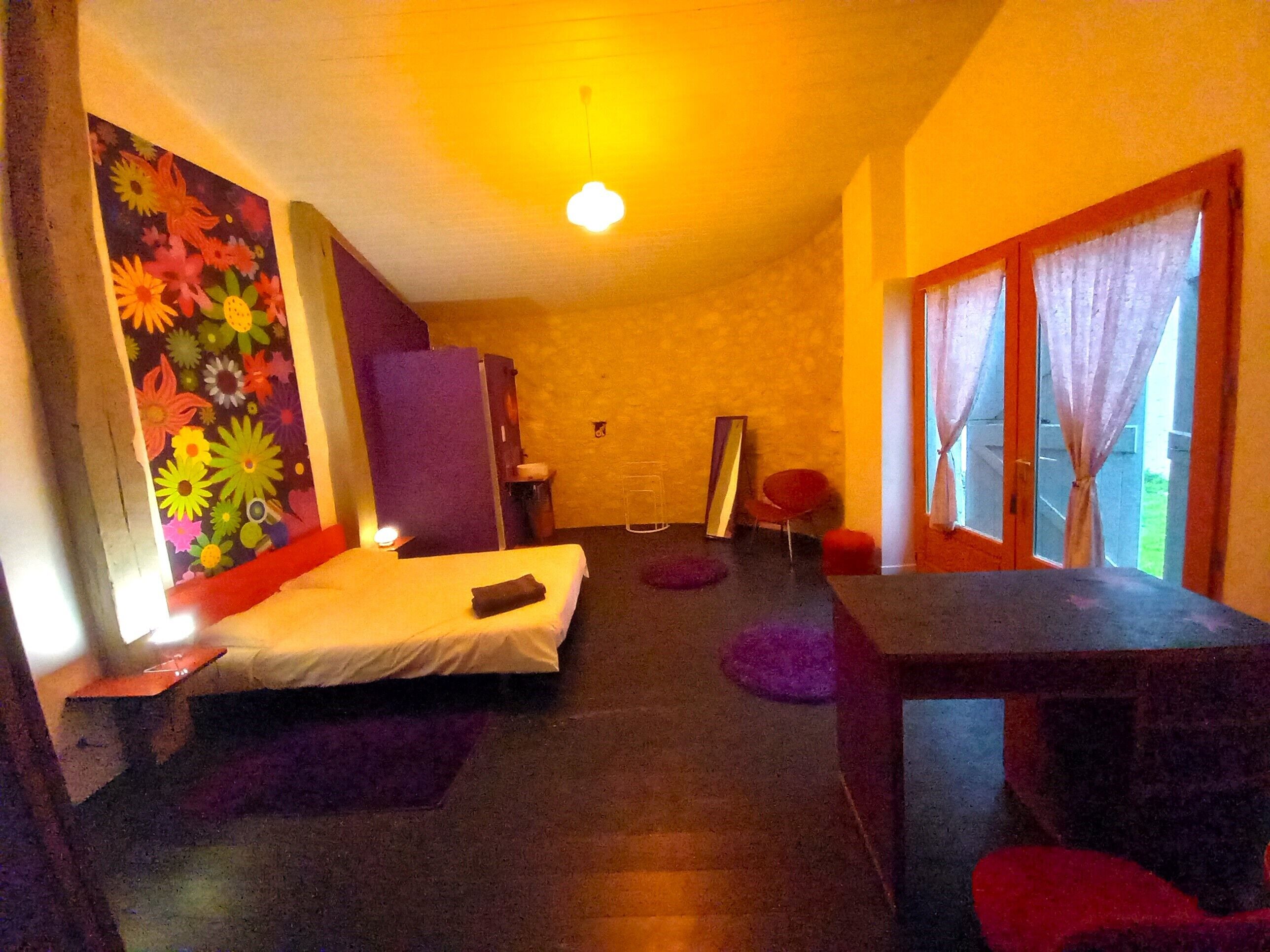 A room with a bed, a desk, a chair, a window with curtains, a wall with a floral mural, and a door.
