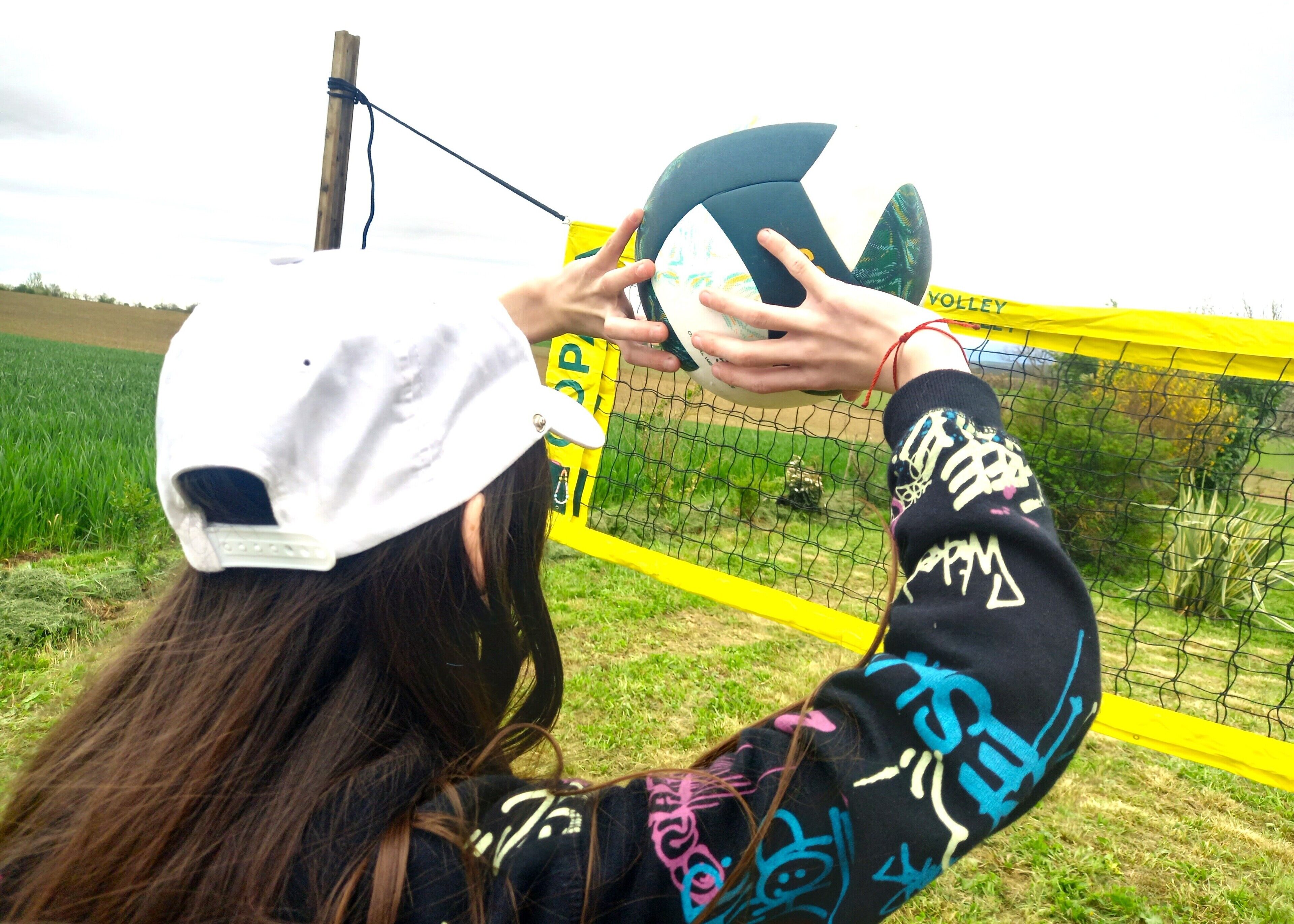 Een persoon houdt een volleybal vast in een grasveld, met een geel net op de achtergrond.