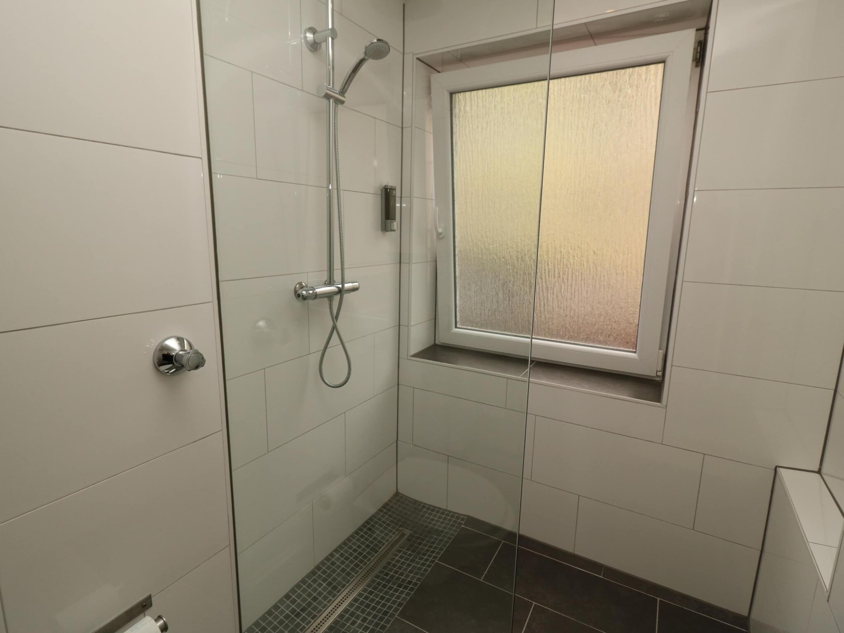 A small, modern bathroom with white tiles, a shower with a handheld showerhead, and a window with frosted glass.