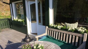 A wooden deck with a round table and two potted plants, a bench with cushions, and a door leading to a covered area with plants. - Holiday Home Waldhaus am Burbach (Bad Berleburg)