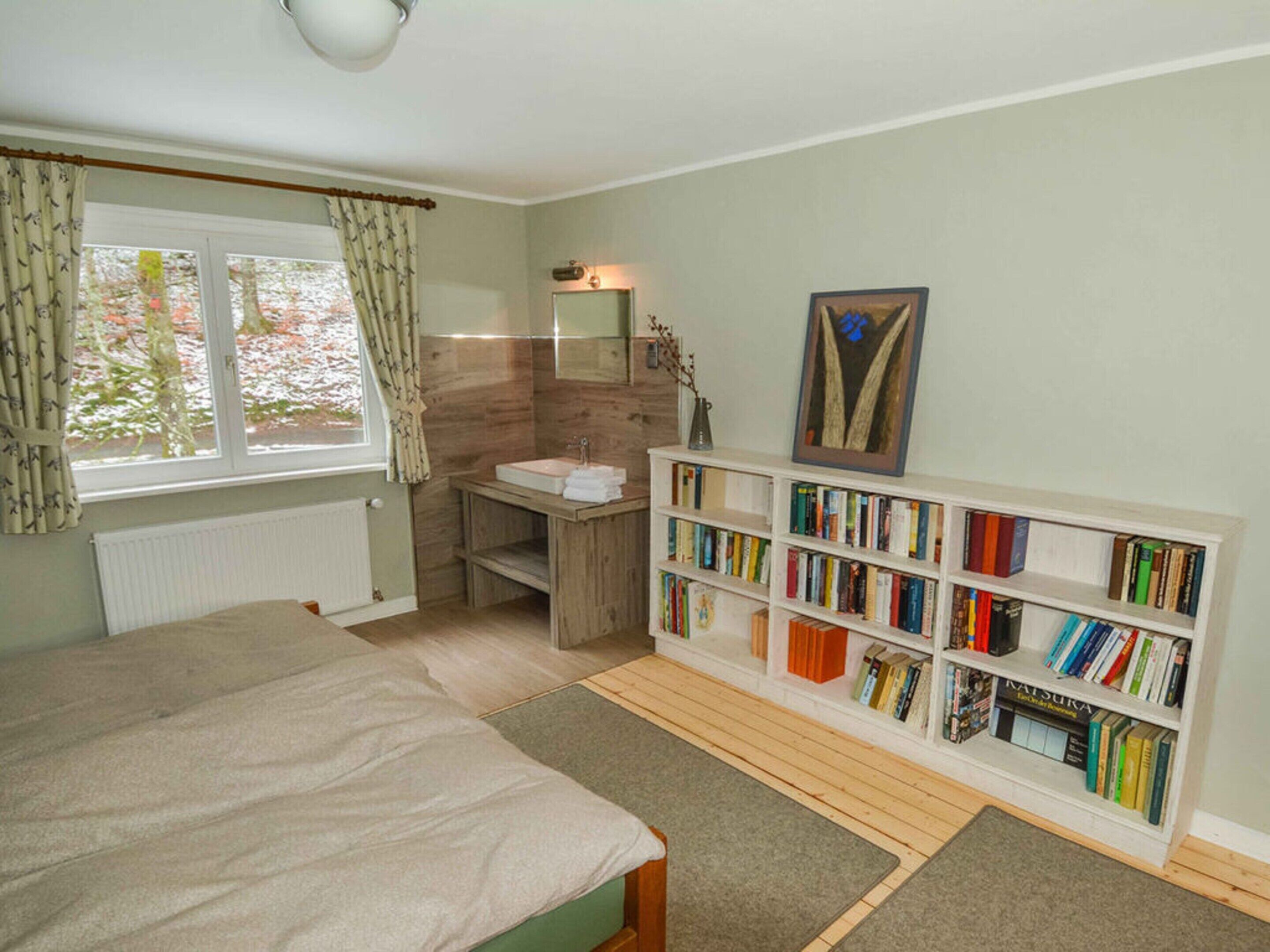 A bedroom with a bed, a bedside table, a bookshelf, a window with curtains, and a bathroom with a sink and mirror.