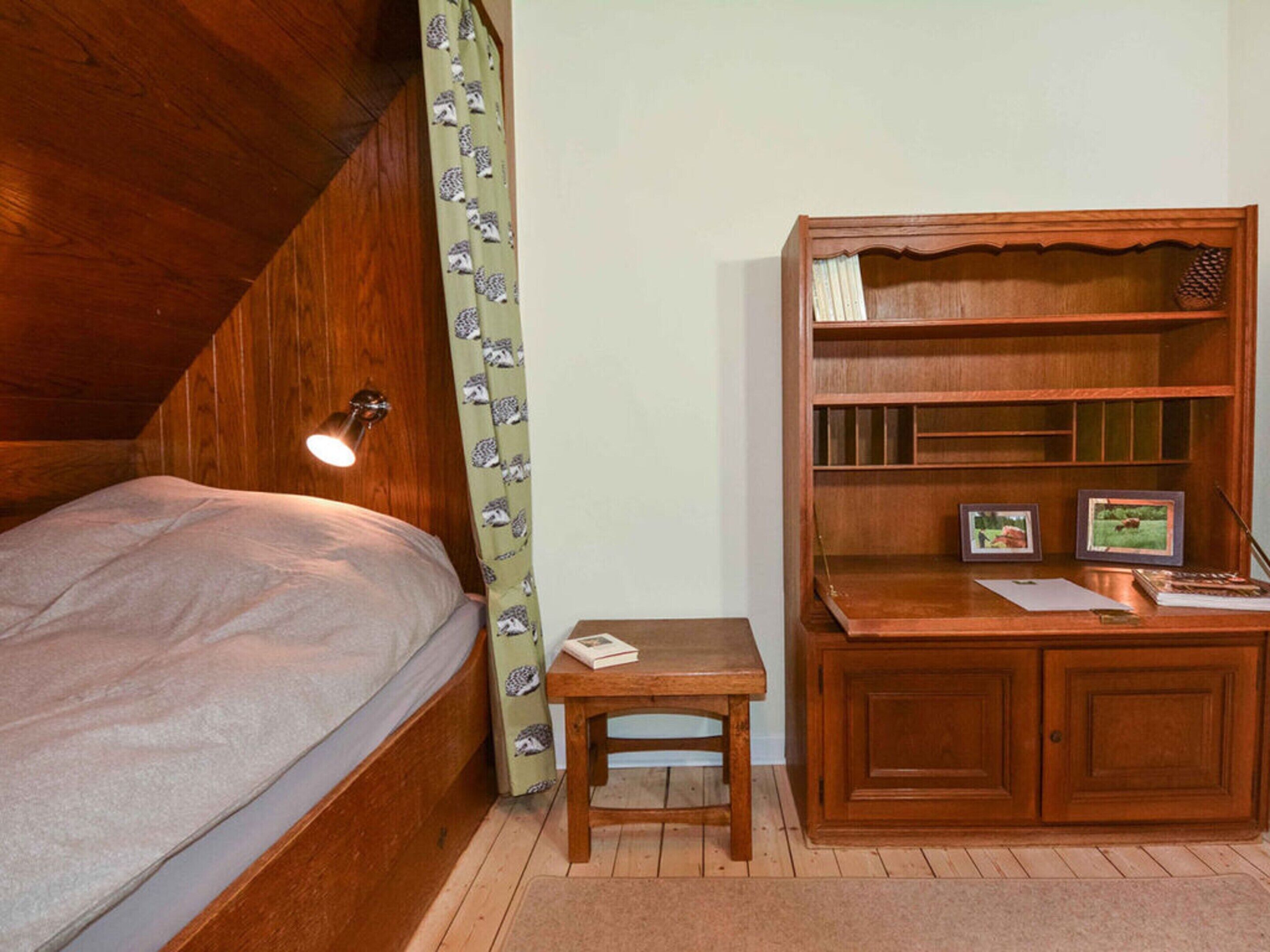 A bedroom with a wooden bunk bed, a bedside table with a lamp, a small wooden desk with a chair, and a wooden bookshelf.