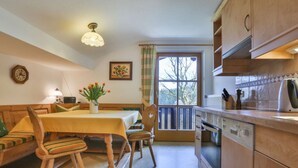 A cozy kitchen with wooden cabinets, a dining table with chairs, a window with curtains, and a clock on the wall. - Haus Brandl Modern Retreat (Ramsau bei Berchtesgaden)