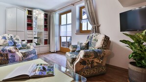 A living room with two floral armchairs, a glass coffee table with a book, a bookshelf, a television, and a large window with curtains. - Haus Brandl Modern Retreat (Ramsau bei Berchtesgaden)