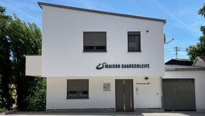 Moderni rakennus, jonka julkisivussa on kyltti, jossa lukee ”MAISON SAARSCHEIFE”. Rakennuksessa on tasakatto, kaksi ikkunaa ja ovi.