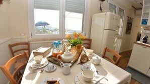 Una mesa de comedor servida con platos, tazas, una tetera y un pastel. Se ve una ventana con persianas, un refrigerador y un reloj en la pared.