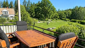 En træterrasse med et bord og stole, der vender ud mod en velplejet græsplæne og træer.