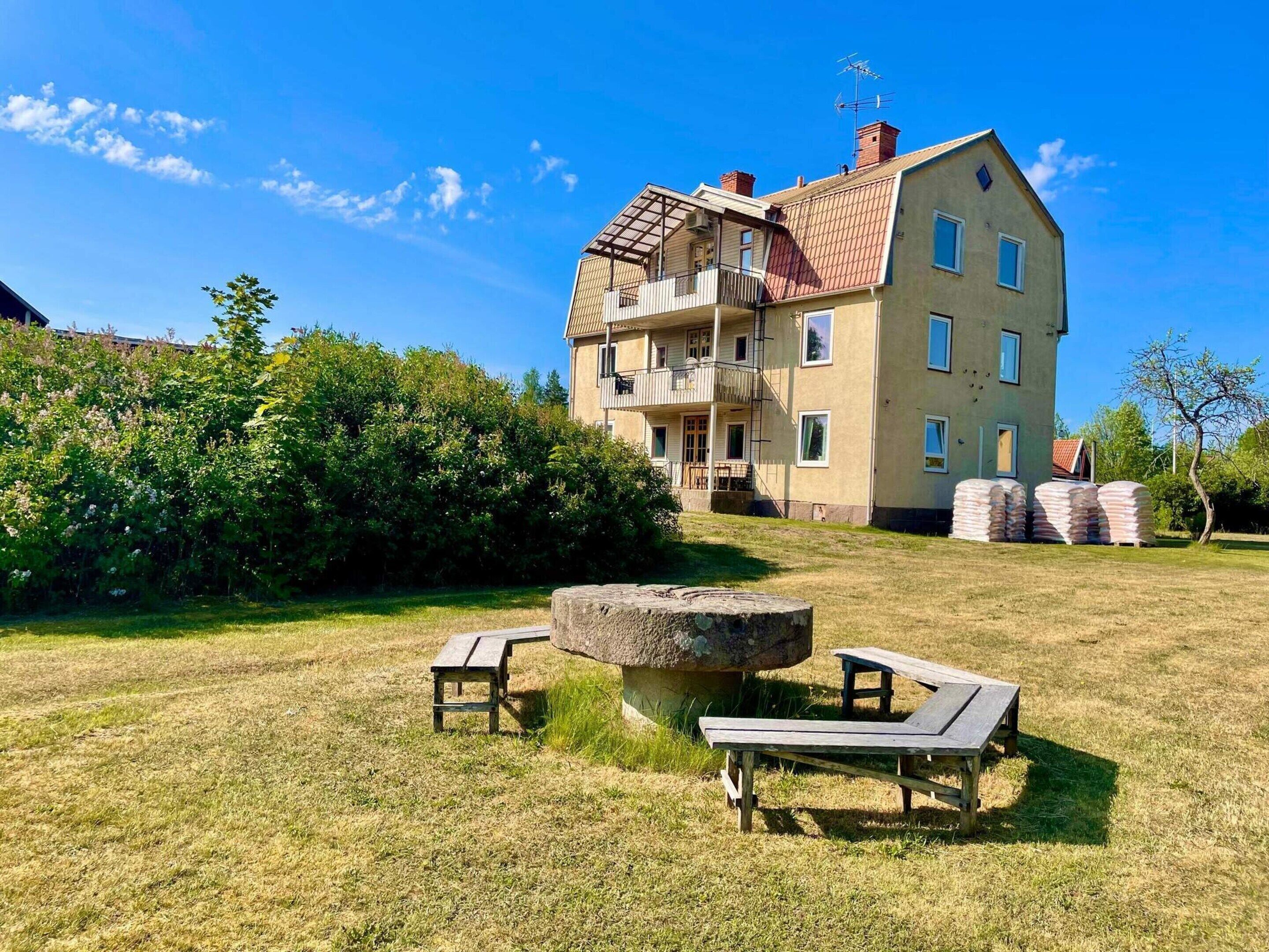A two-story building with a balcony, a stone table, and a picnic table in a grassy area.