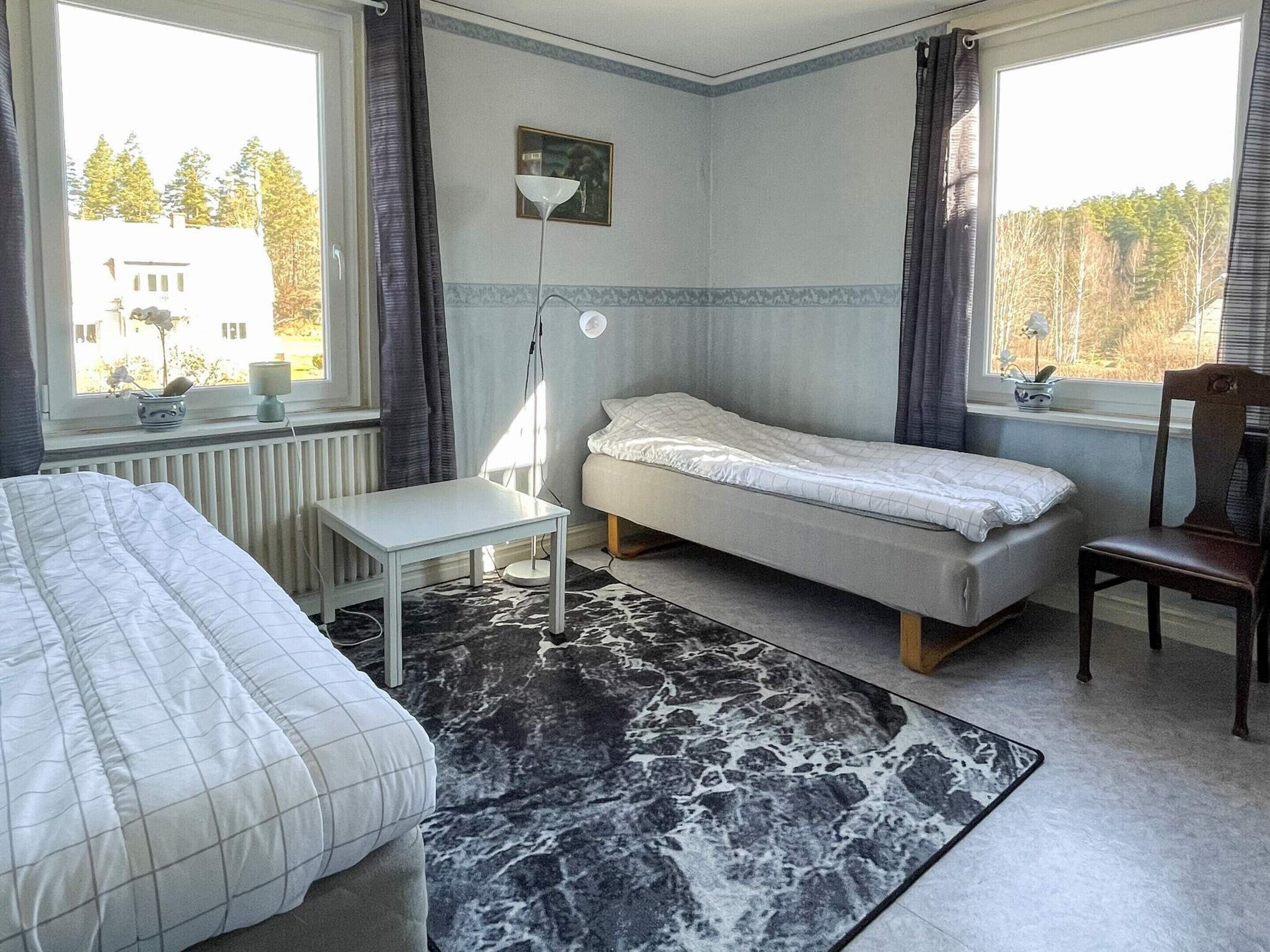 A bedroom with a bed, a small table, a chair, a floor lamp, and a view of trees through the window.