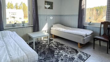 A bedroom with a bed, a small table, a chair, a floor lamp, and a view of trees through the window.