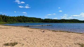 A sandy beach with a dock extending into a calm lake, surrounded by dense forest.