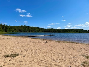 En sandstrand med en brygga som sträcker sig ut i en lugn sjö, omgiven av tät skog.