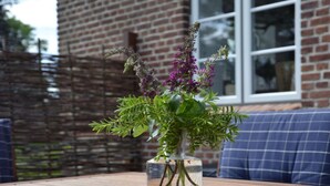Un vase en verre transparent, orné de feuillage vert et de fleurs violettes, est posé sur une table en bois.