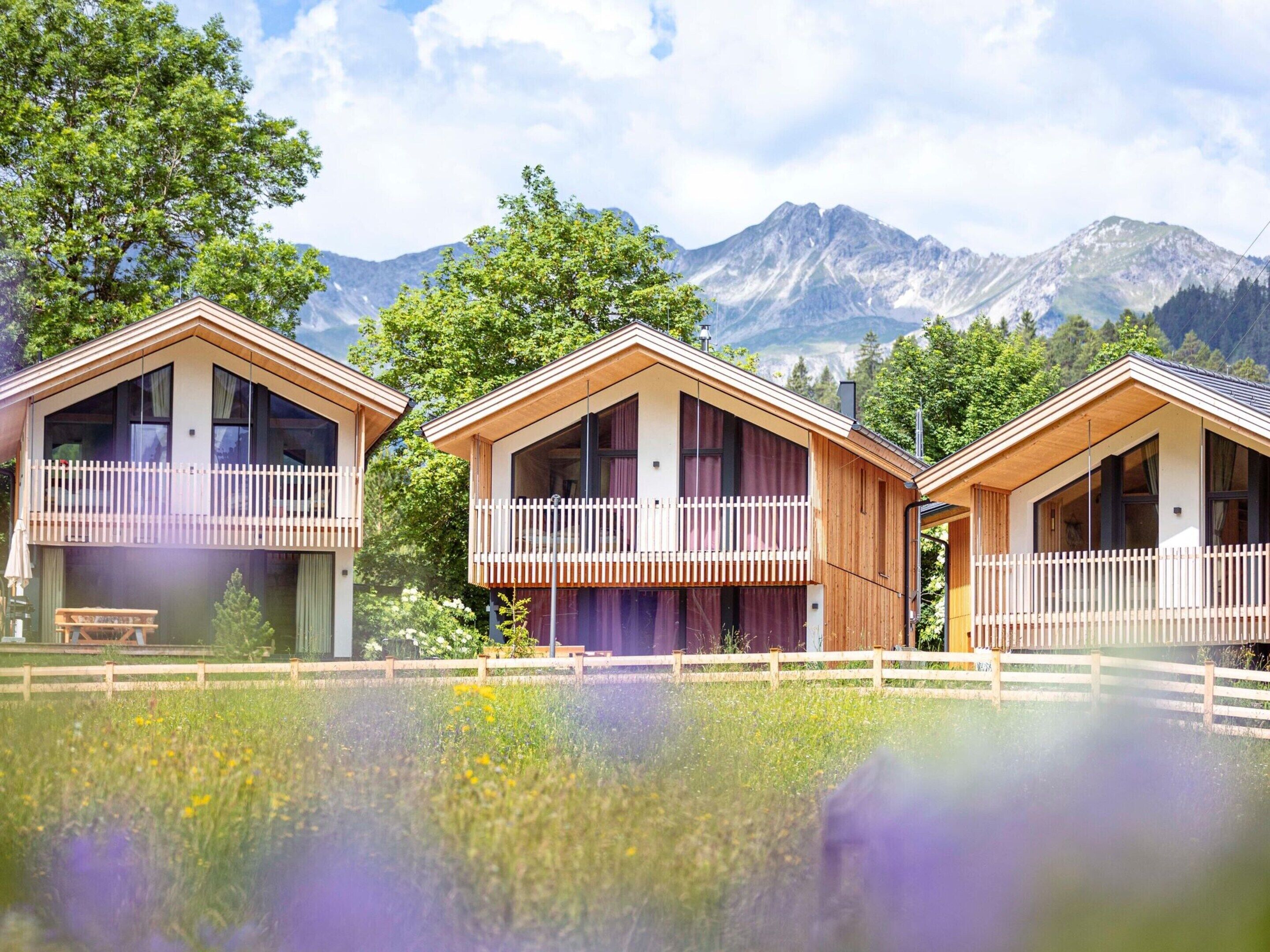 Three wooden cabins with balconies, set in a meadow with mountains in the background.