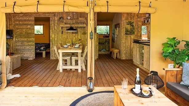 A rustic cabin interior with a wooden dining table, a kitchen area, and a seating area with a cushioned bench. - Beautiful Glamping Tent With Pellet Stove (Winterswijk - Brinkheurne)