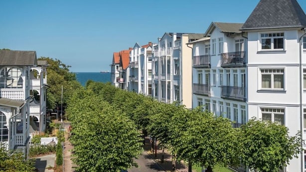 A row of modern multi-story residential buildings with balconies, a clear blue sky, and a view of the sea in the background. - "seashell" Modern Retreat (Binz)