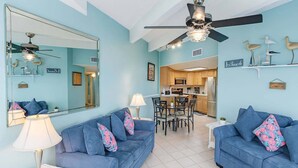 A living room with two blue sofas, a dining table, and a kitchen area. - Aquarius 408 2 Bedroom Condo by RedAwning (South Padre Island)