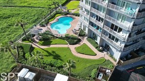 An aerial view of a hotel with a swimming pool, lush green lawns, and a multi-story building with balconies. - Aquarius 408 2 Bedroom Condo by RedAwning (South Padre Island)