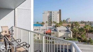 A balcony with a view of a building complex, including a tall structure with a unique design and a smaller building with a red roof. - Aquarius 408 2 Bedroom Condo by RedAwning (South Padre Island)
