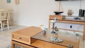 A modern living room with a glass coffee table, wooden side table, and a dining area with chairs and a table. - Beach & Palms Condo 2 Bedroom Condo by RedAwning (South Padre Island)