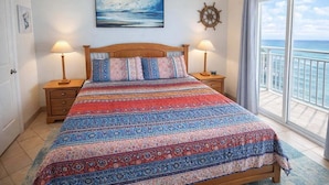 Un dormitorio con una cama grande, dos mesitas de noche, un cuadro, una decoración con timón de barco y vistas al océano a través de una puerta corredera.