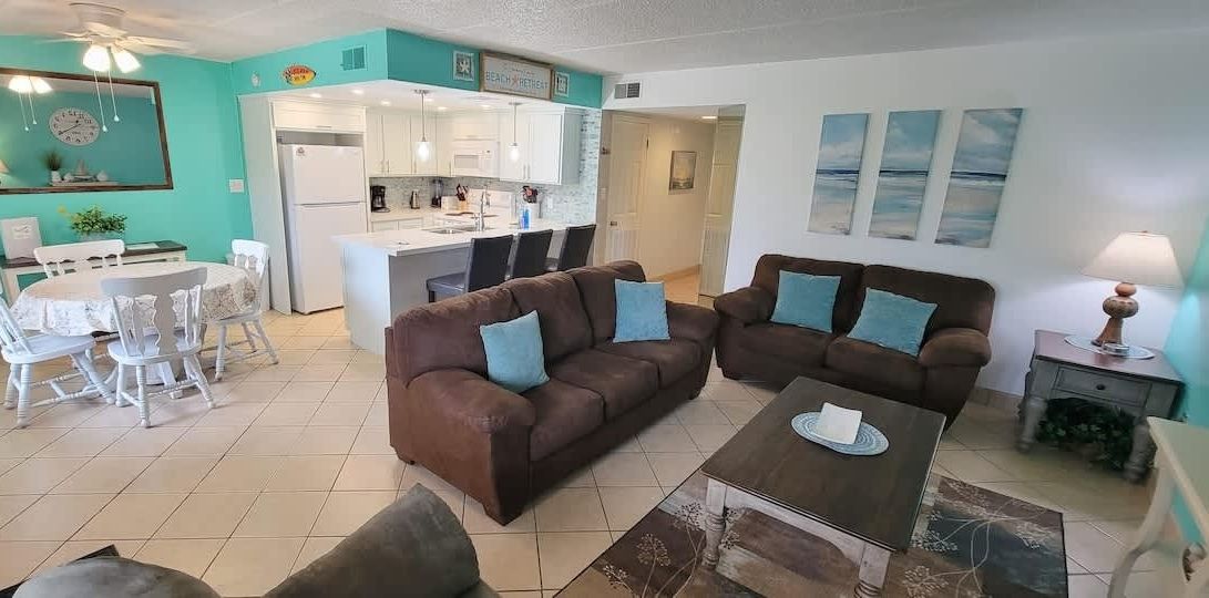 A living room with a brown sofa, a dining table with white chairs, and a kitchen in the background.