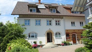 A two-story building with a tiled roof, a central entrance with an archway, and a sign above the entrance. - Apartment in Hergensweiler (Hergensweiler)