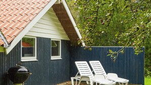 Ein Haus mit überdachter Terrasse, Grill und zwei weißen Lounge-Stühlen.
