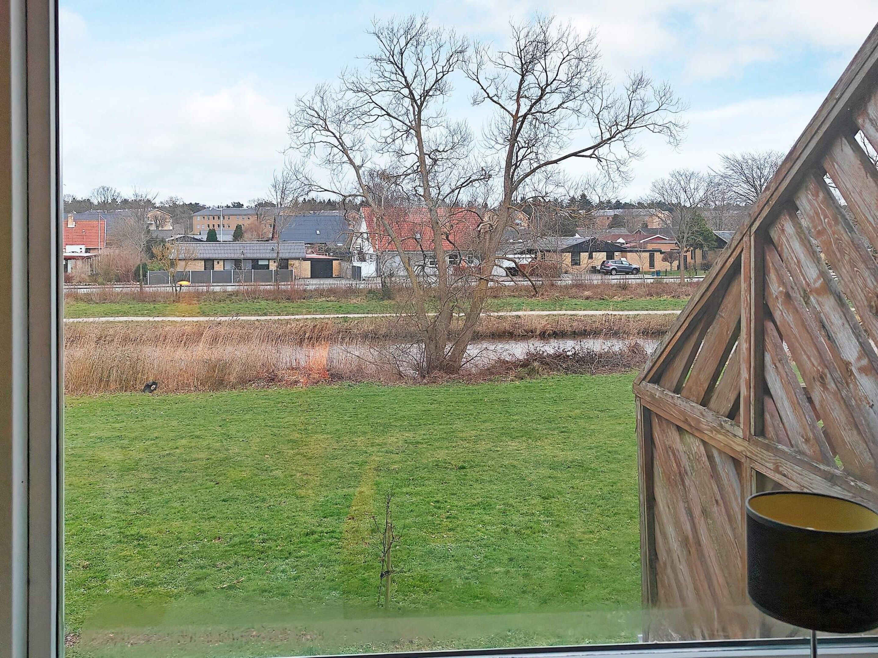 Une vue à travers une fenêtre, avec une structure en bois, un champ herbeux et des maisons en arrière-plan.