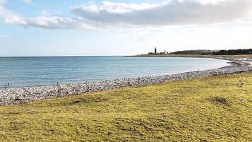 Un paisaje costero con una zona cubierta de hierba, una playa de guijarros y un faro a lo lejos.