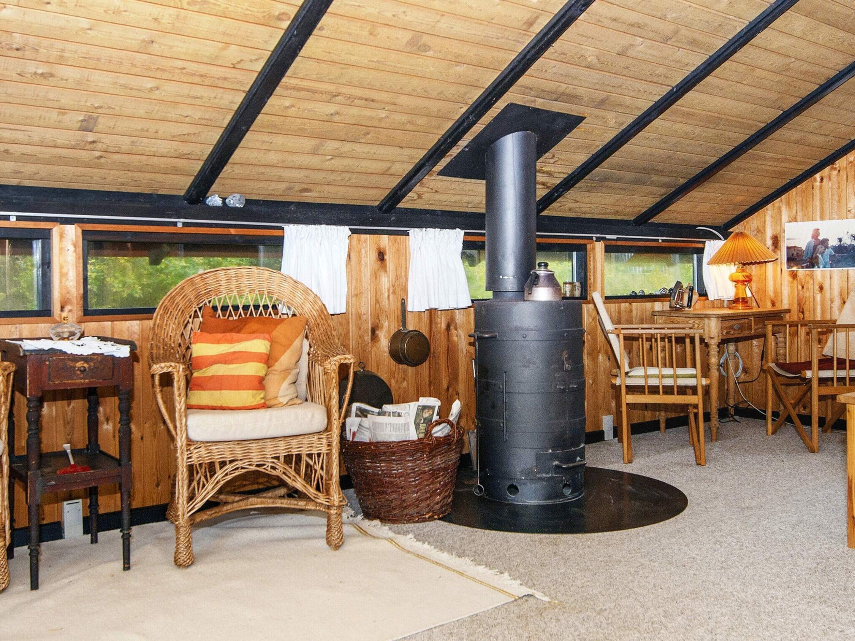 A cozy room with a wooden ceiling, a wicker chair with a cushion, a table with chairs, and a black stove.
