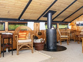 Una habitación acogedora con techo de madera, una silla de mimbre con cojín, una mesa con sillas y una estufa negra.