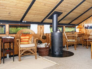 Une pièce chaleureuse avec un plafond en bois, un fauteuil en osier avec un coussin, une table avec des chaises et un poêle noir.
