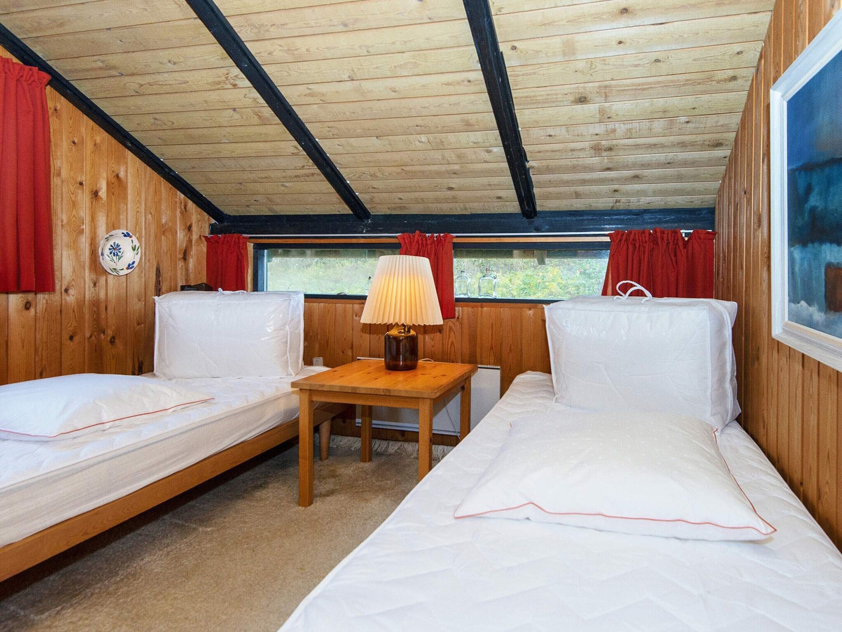 Una cabaña con dos camas individuales, una mesa pequeña con lámpara, paredes de madera y una ventana con cortinas.